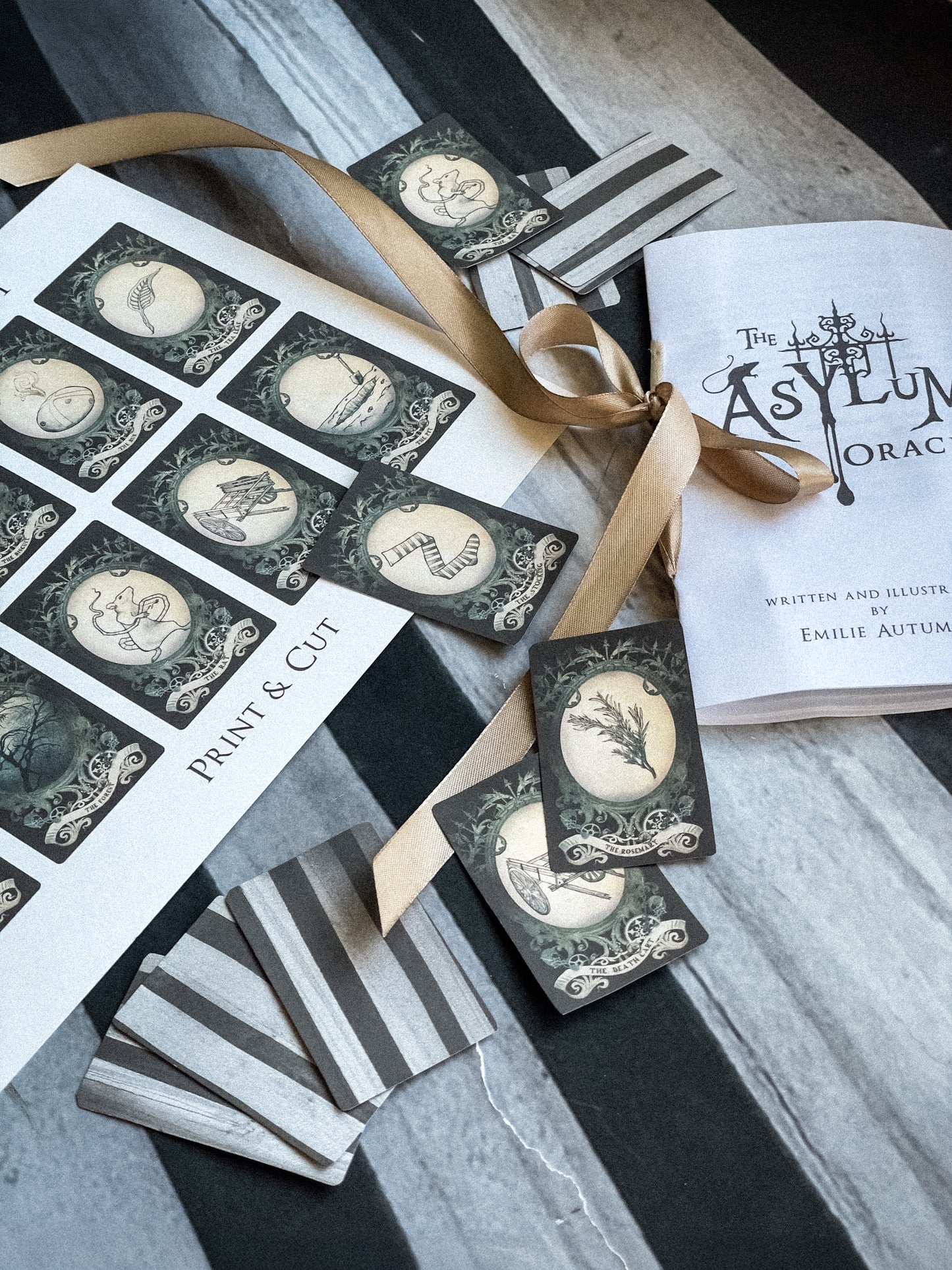 Collection of printed cards with decorative designs and a book titled 'The Asylum Oracle' on a striped surface.