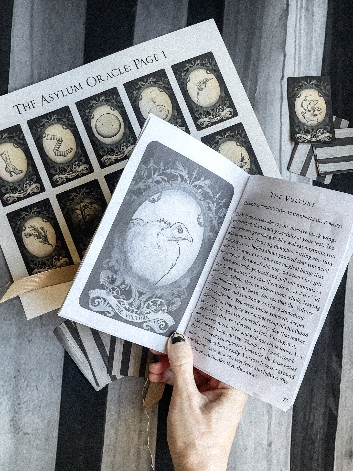 Open book with 'The Asylum Oracle' cards displayed on a striped fabric background